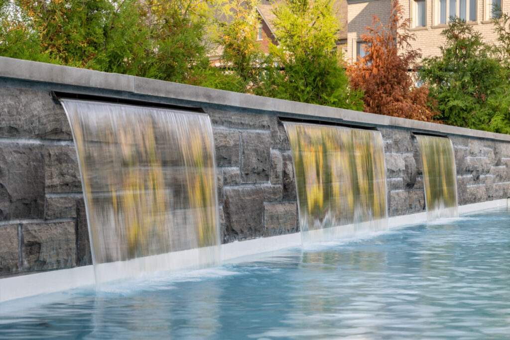 The image shows a serene water feature with four smooth waterfalls cascading into a clear pool, backed by a stone wall, with lush greenery around.