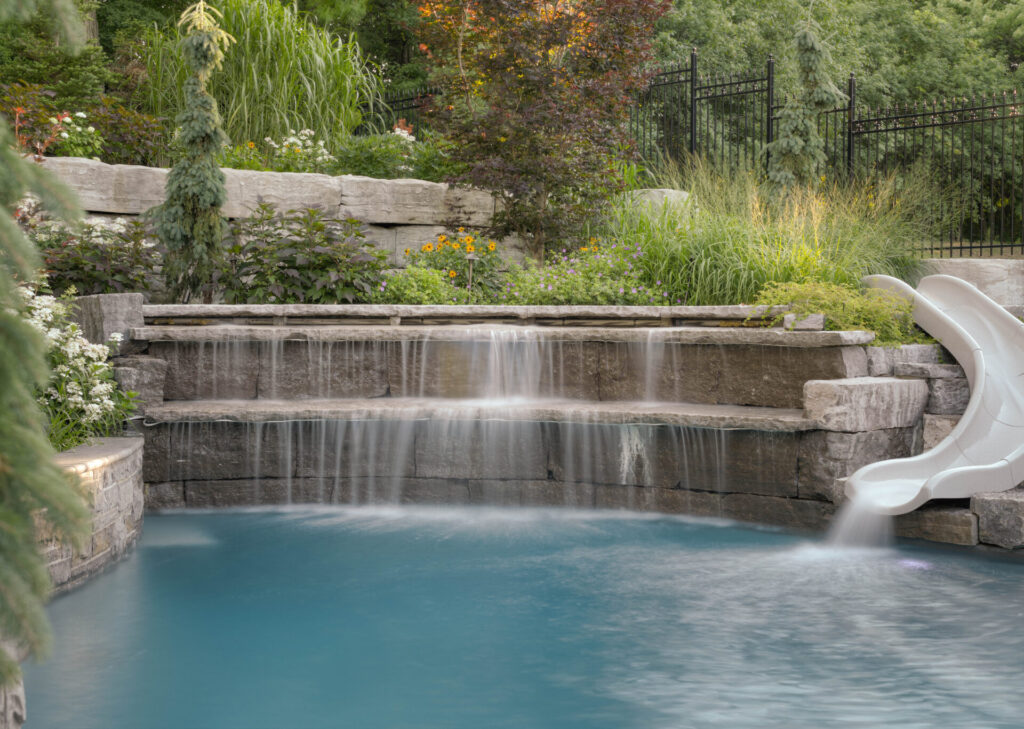 An idyllic backyard with a cascading stone waterfall into a pool, lush greenery, a white slide, and a wrought iron fence under a soft light.