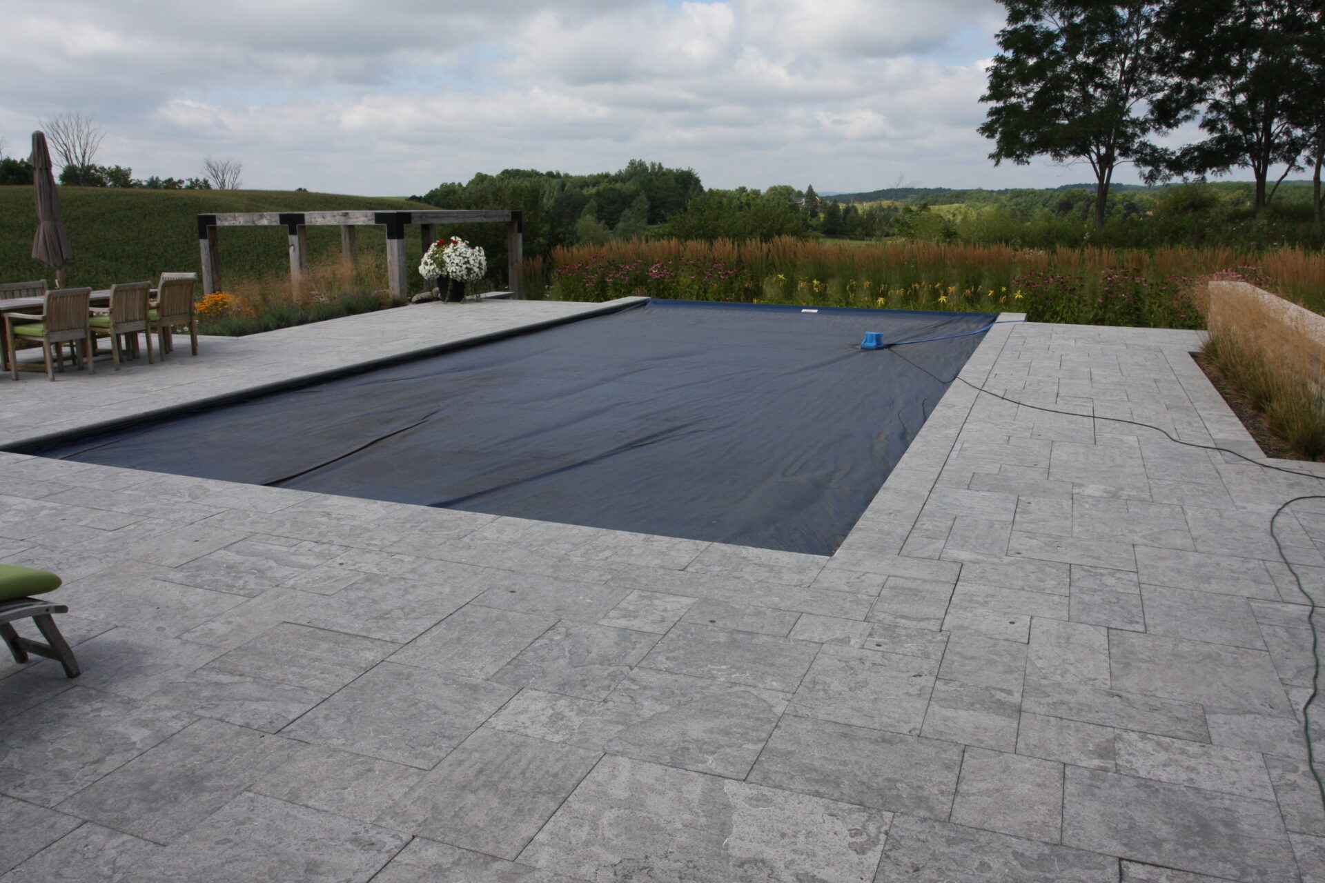 An outdoor swimming pool covered with a dark tarp, surrounded by patio and furniture, overlooking a natural landscape with trees and fields.