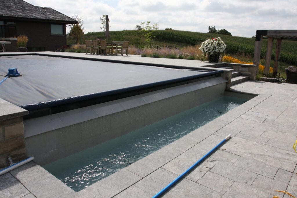 This image shows a rectangular swimming pool partially covered with a grey safety cover, alongside patio furniture and landscaped garden in the background.