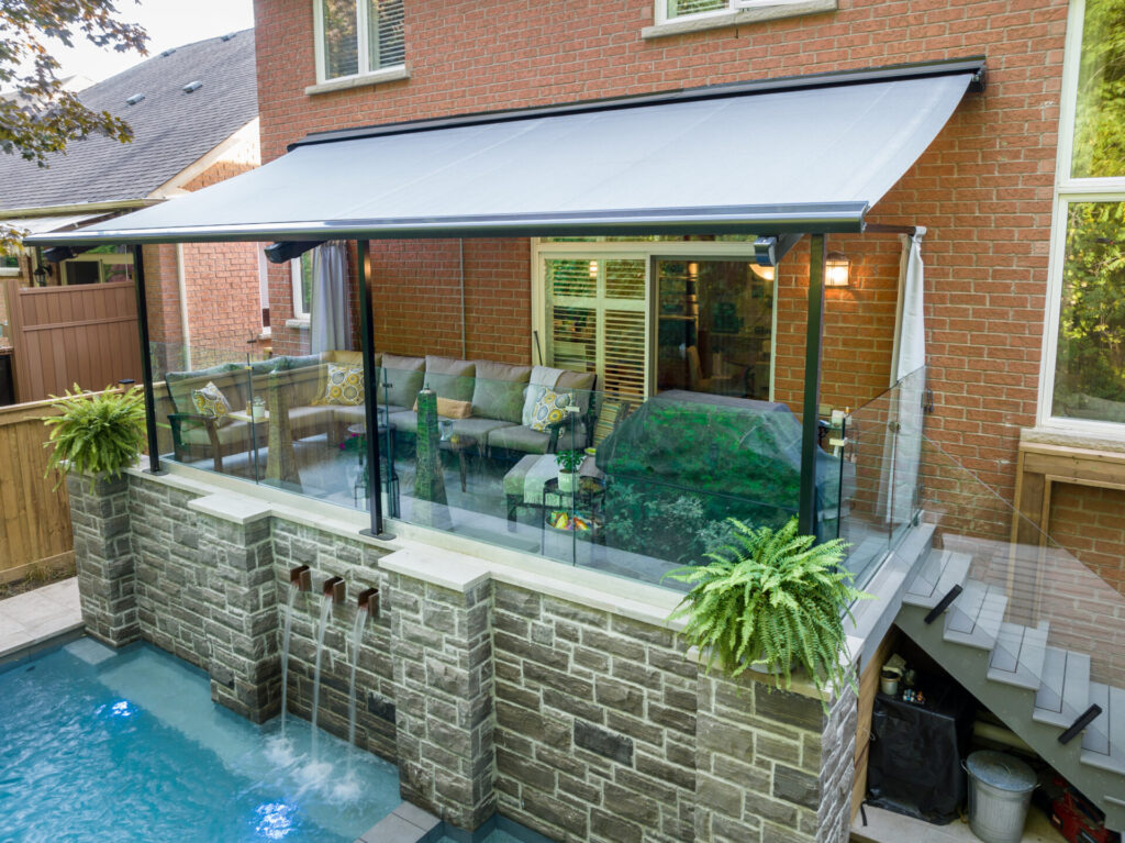 A modern outdoor patio with a retractable awning overlooks a swimming pool with water features, encased by a brick house and glass railings.
