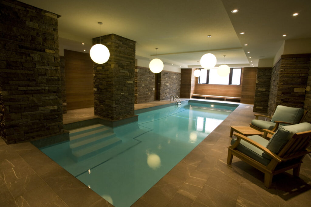 An indoor swimming pool with clear blue water, surrounded by stone pillars and walls. Wooden lounge chairs sit by the poolside under spherical lights.