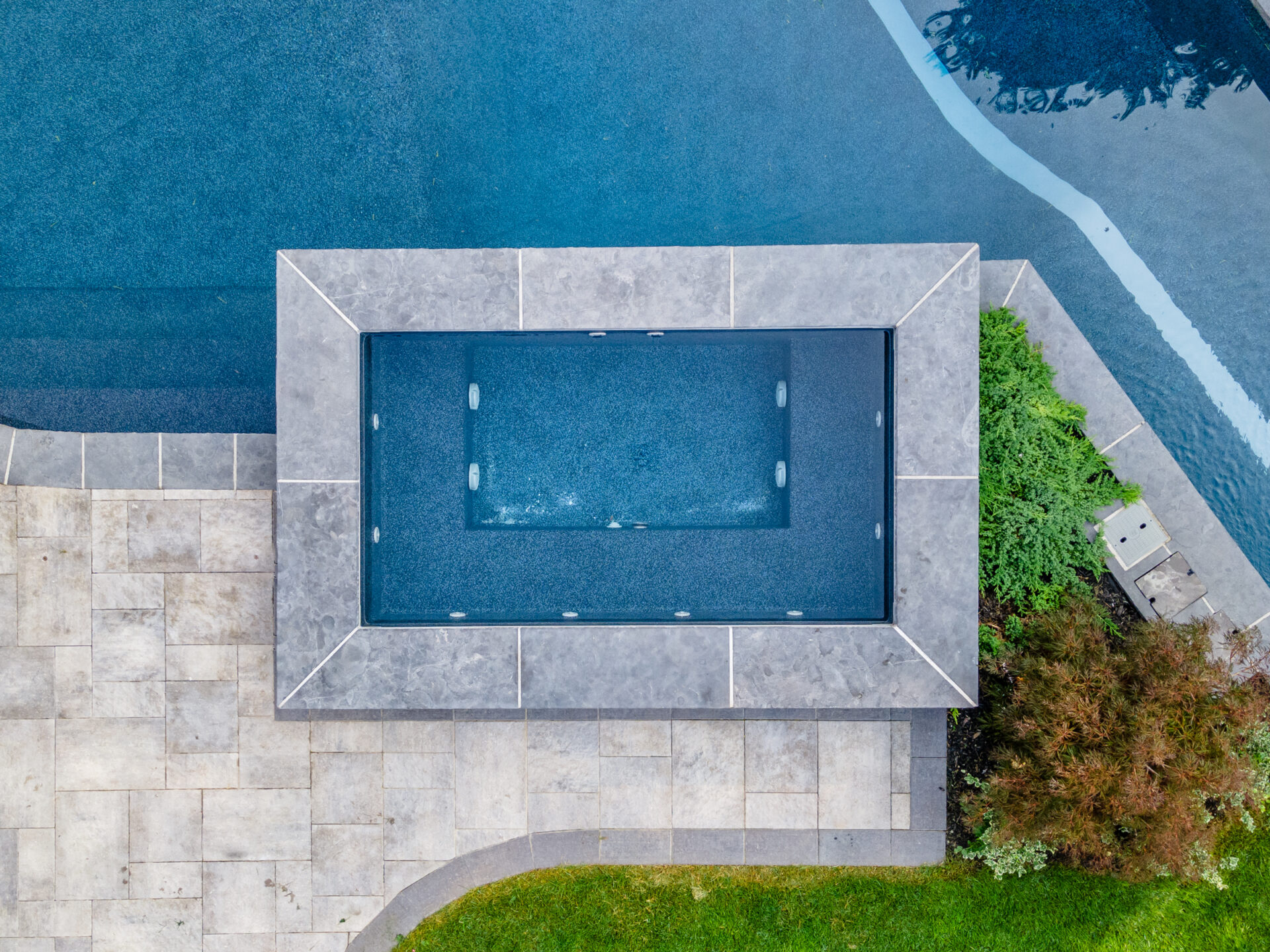 An aerial view of a backyard with a rectangular swimming pool surrounded by tiled decking, adjacent to a larger body of water, with landscaping.