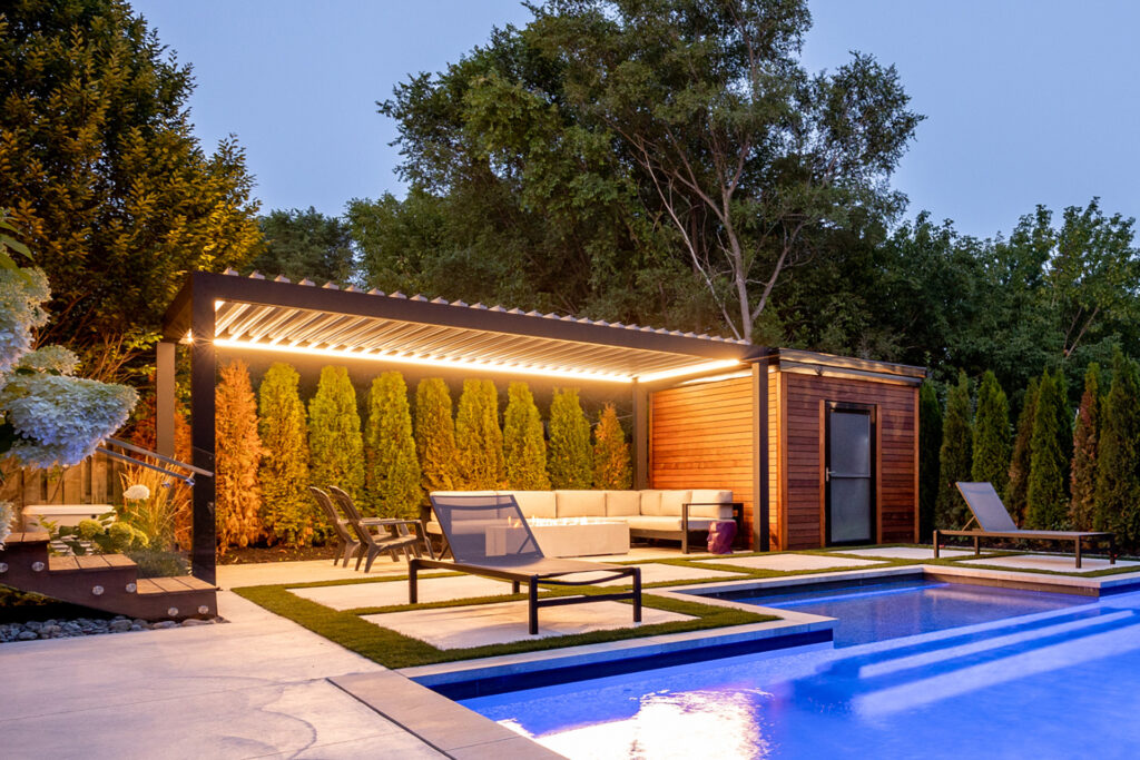 A modern backyard at dusk features a lit pergola, outdoor seating, lush greenery, a swimming pool with a sun lounger, and a cozy ambiance.