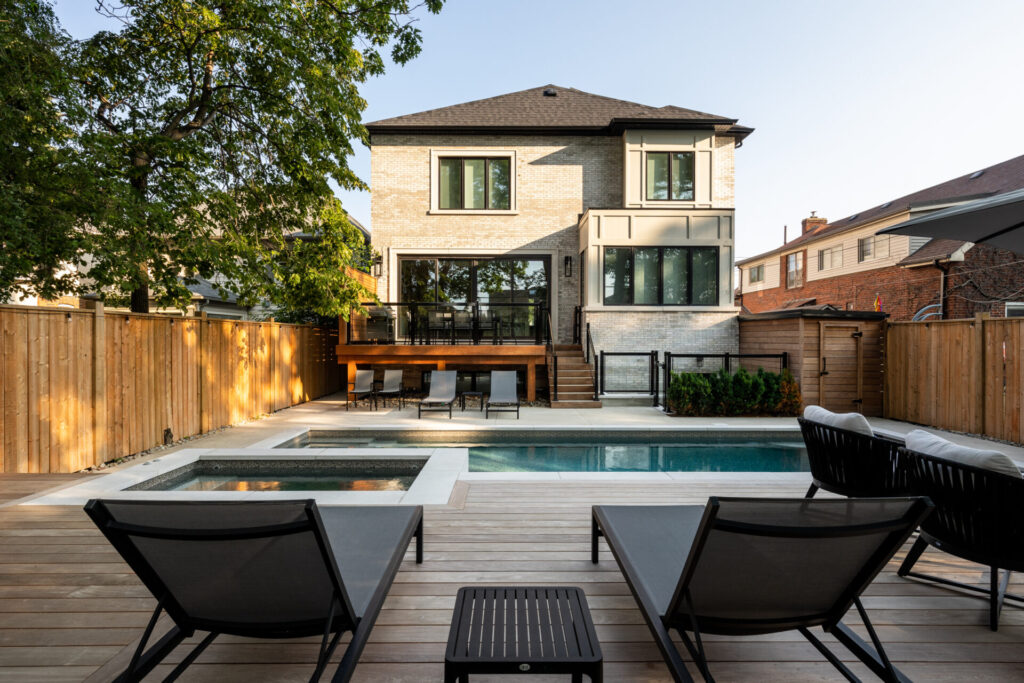 A modern two-story house with a pool, hot tub, and wooden deck, featuring outdoor furniture, a fence, and adjacent neighborhood homes in view.