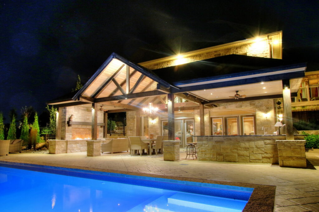 An outdoor luxury patio with a fireplace, seating, and bar area next to an illuminated pool during nighttime, showcasing a relaxed entertainment space.
