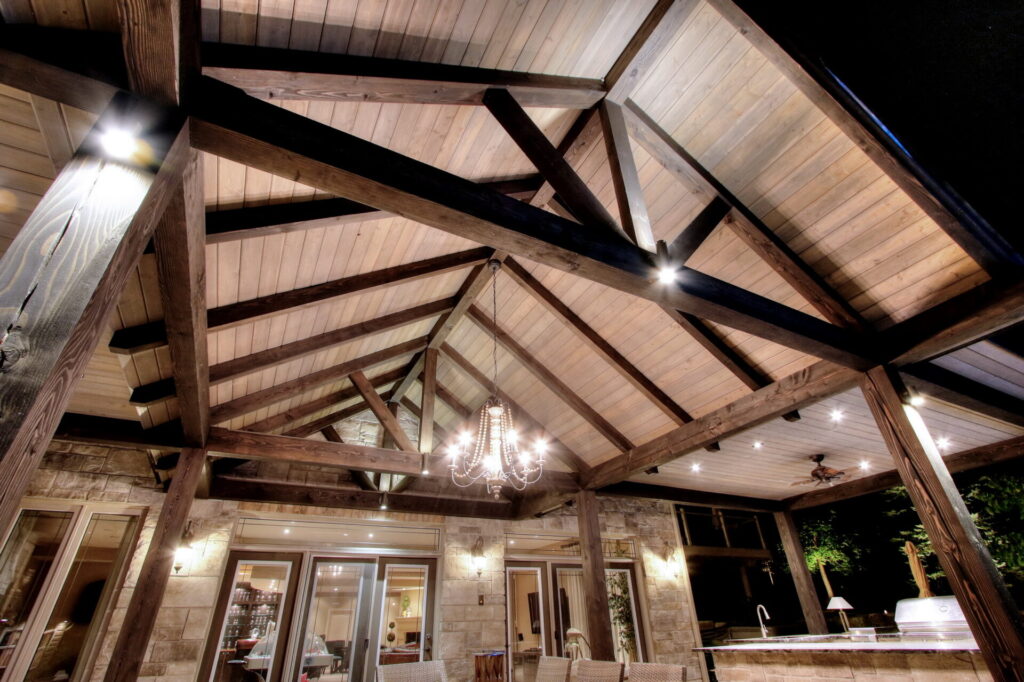 Elegant outdoor covered patio with wooden exposed beams, ceiling fans, and chandelier, leading into a warmly lit interior through glass doors.