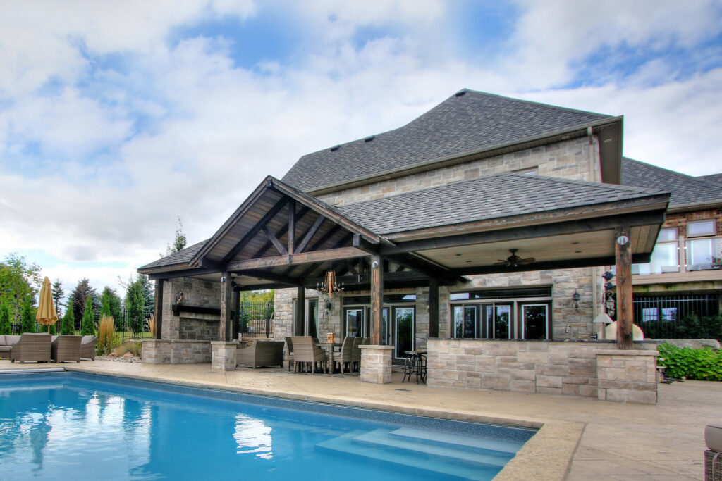 Luxurious house with a pool, outdoor patio, lounge chairs, and stone fireplace. The architecture features a tiled roof and large windows set in a tranquil environment.
