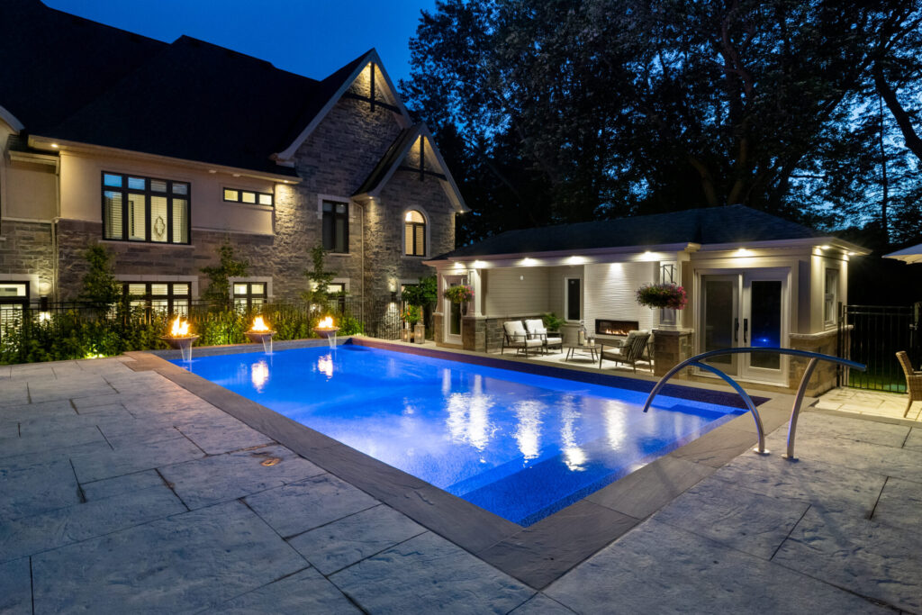 The image shows an illuminated backyard with a swimming pool at dusk, a stone house facade, patio furniture, and torch flames beside the pool.