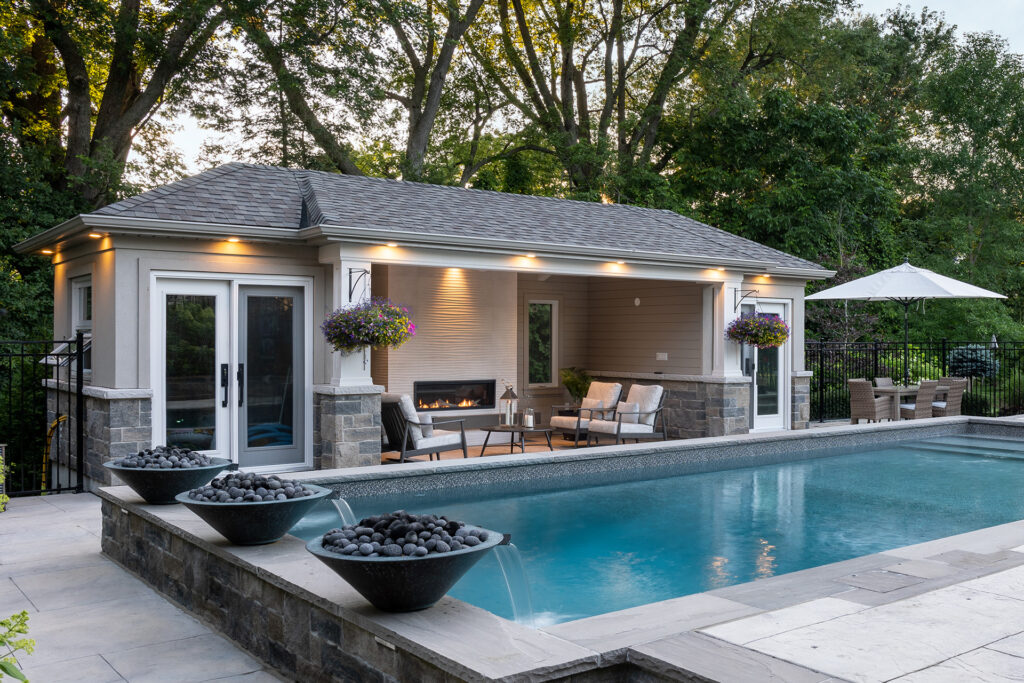 This image shows an elegant backyard with a swimming pool, a pool house, comfortable outdoor seating, decorative plants, and evening ambient lighting.