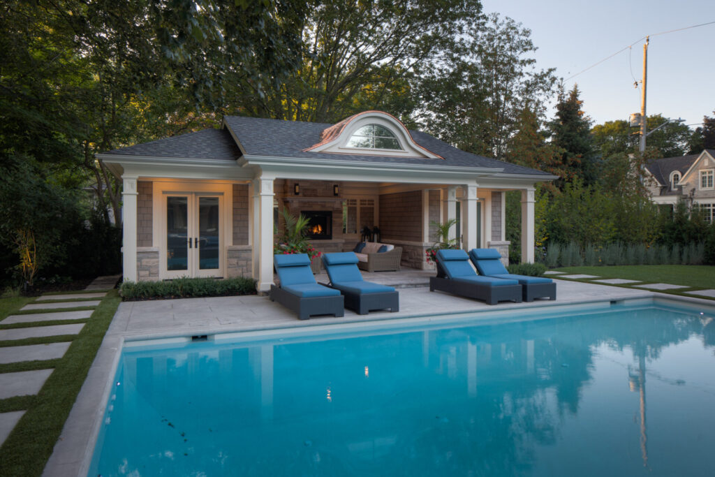 Elegant poolside area during twilight with a swimming pool, sun loungers, a covered patio area, landscaped garden, and a pool house with lighting inside.