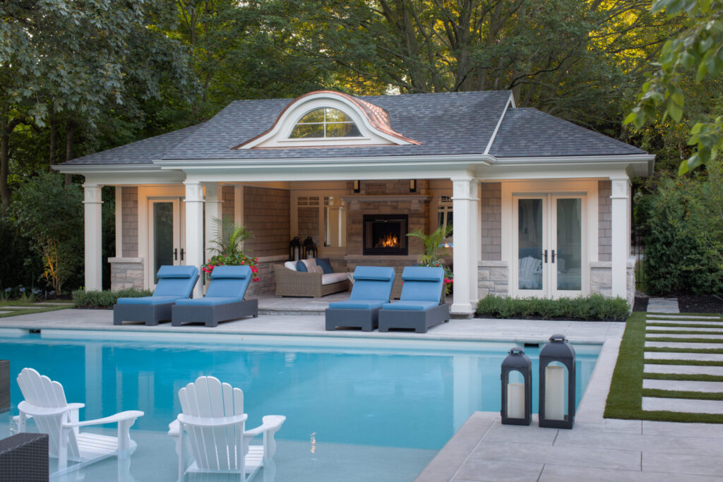 An elegant pool house with a fireplace overlooks a serene swimming pool, flanked by comfy loungers and classic white Adirondack chairs in a lush backyard.