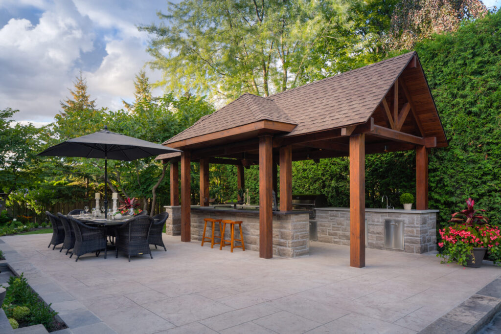 An outdoor patio with a dining set under an umbrella, a wooden pavilion with a bar counter, and lush greenery in the background.