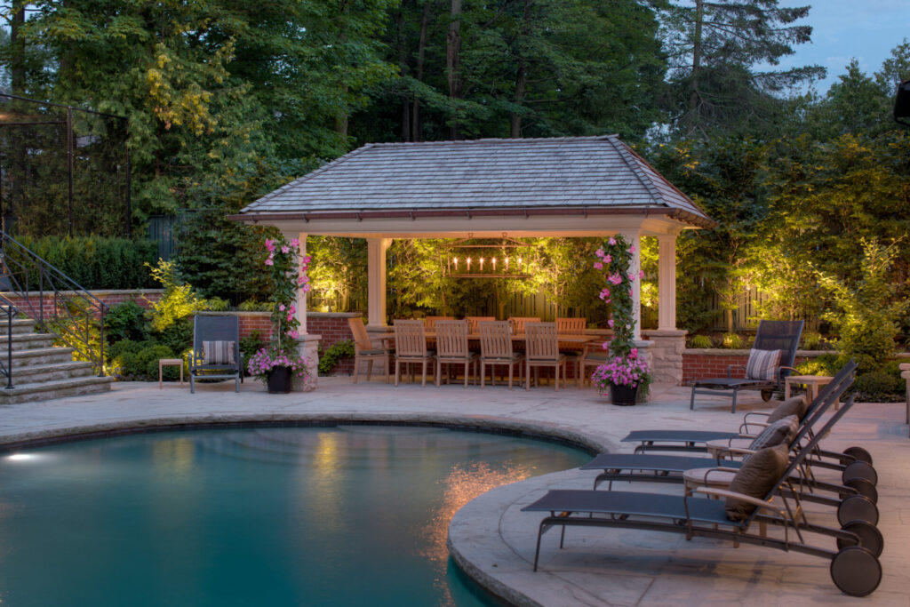 An elegant poolside evening setting featuring a pergola adorned with lights, inviting chairs, blooming flowers, and loungers. A tranquil atmosphere with surrounding greenery.