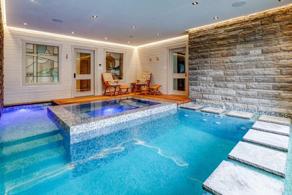 An indoor pool with blue mosaic tiles, featuring stepping stones, flanked by a stone wall on one side and a house entrance on the other.