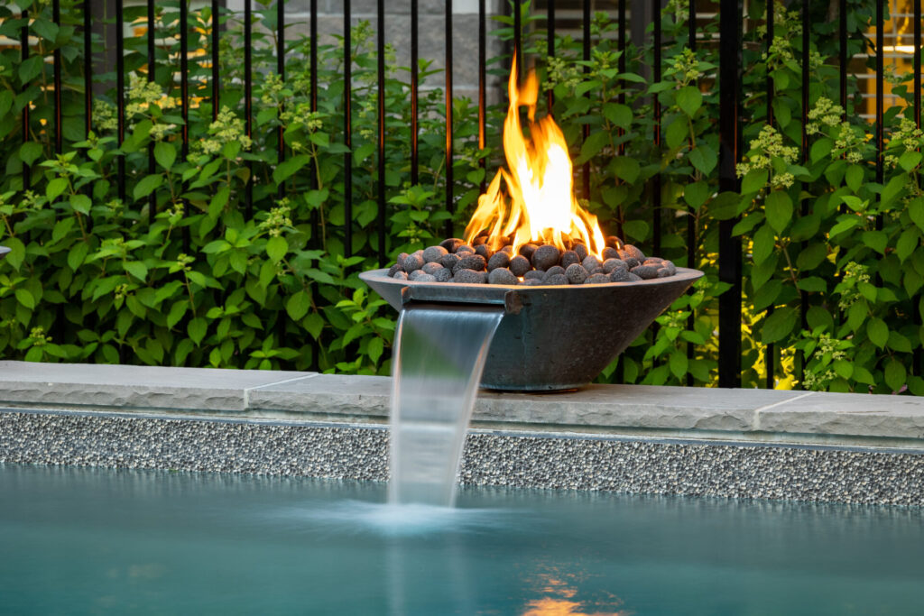 An outdoor fire bowl with flames beside a swimming pool with a smooth waterfall feature, surrounded by dense green foliage and a metal fence.