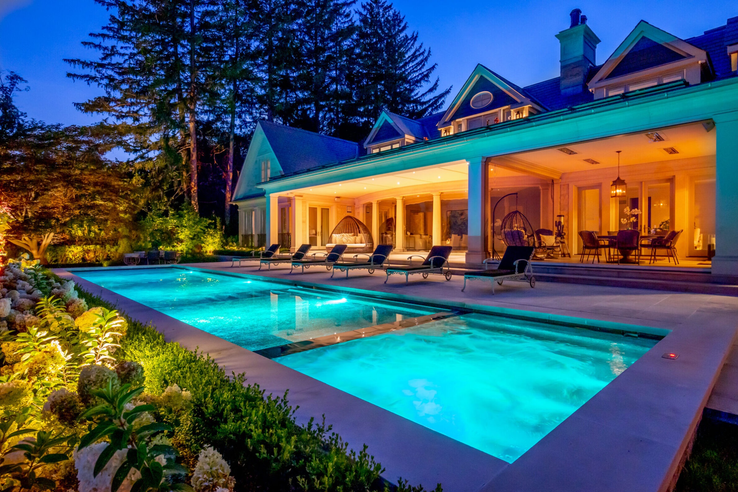 A luxurious house at twilight with illuminated windows, outdoor swimming pool with turquoise water, patio furniture, and surrounded by green landscaping.