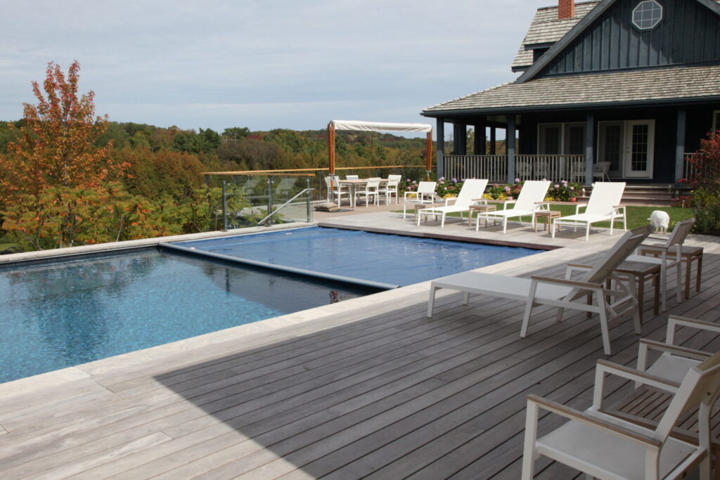 A serene outdoor setting with a clear blue swimming pool, deck chairs, a wooden deck, and a house with a porch set against an autumnal landscape.