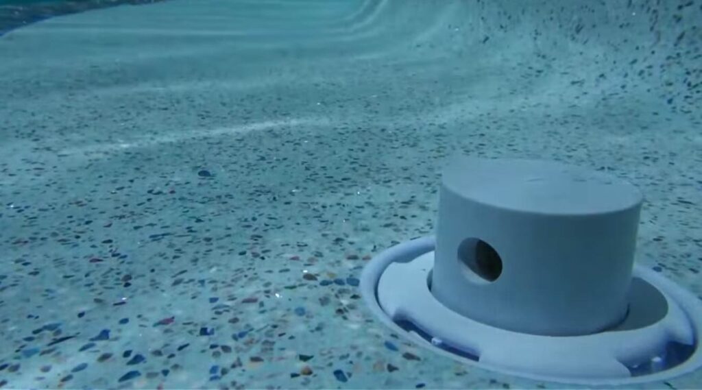 Underwater view of a swimming pool with a main drain cover on the floor, showing pebble aggregate finish and clear blue water.