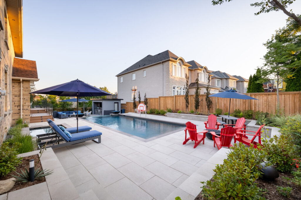 A residential backyard with a rectangular pool, loungers, red Adirondack chairs, a large umbrella, well-kept garden, and a backdrop of elegant houses during twilight.