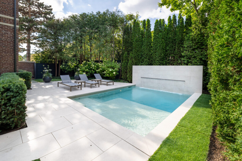 An outdoor residential area featuring a rectangular swimming pool, lounge chairs, manicured greenery, and a privacy hedge under a clear sky.