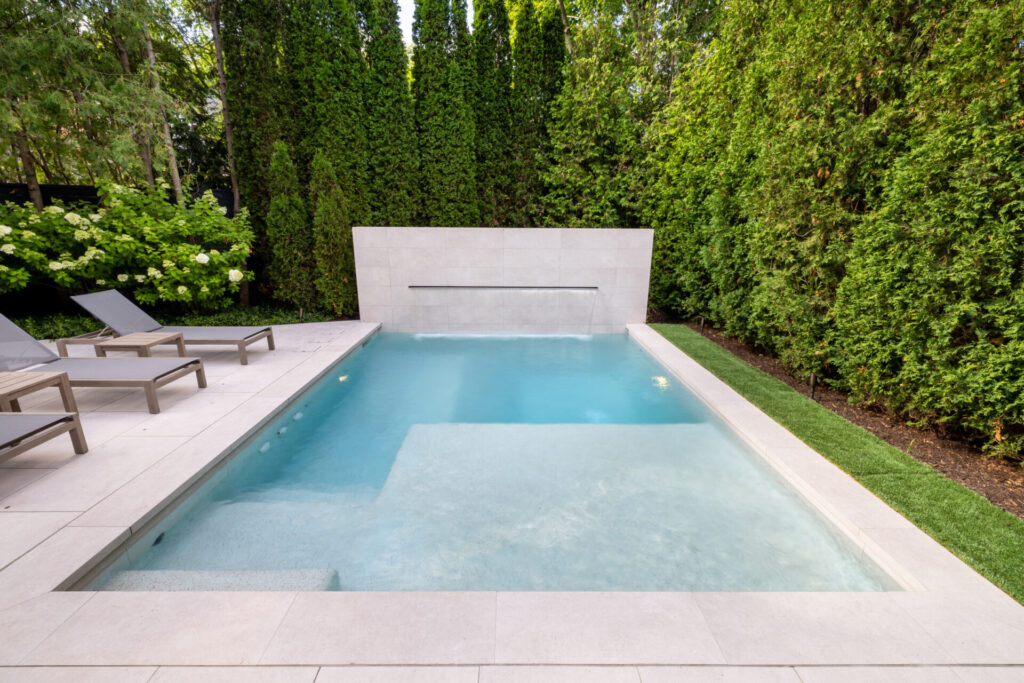 Modern rectangular swimming pool with clear blue water bordered by light tiles, sun loungers, and lush green privacy hedges in a garden.