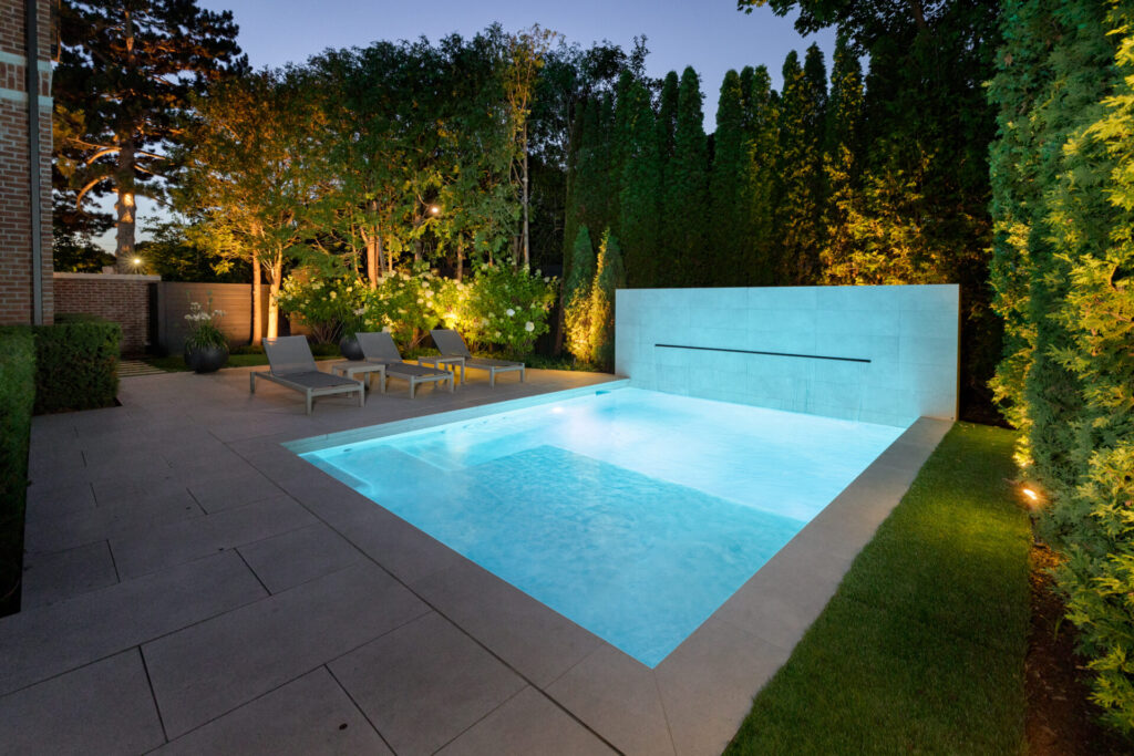 A serene evening setting with a rectangular swimming pool, surrounded by patio loungers, tall privacy hedges, and soft landscape lighting.
