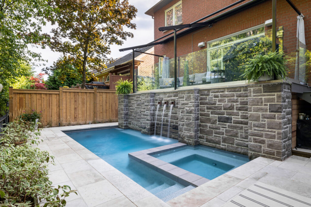 This image shows a backyard with a swimming pool and hot tub. The area is surrounded by a wood fence, stone walls, and a brick house with large windows.