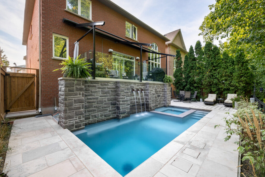 This image shows a rectangular swimming pool in a backyard with stone walls, adjacent to a brick house with large windows, surrounded by lush greenery.