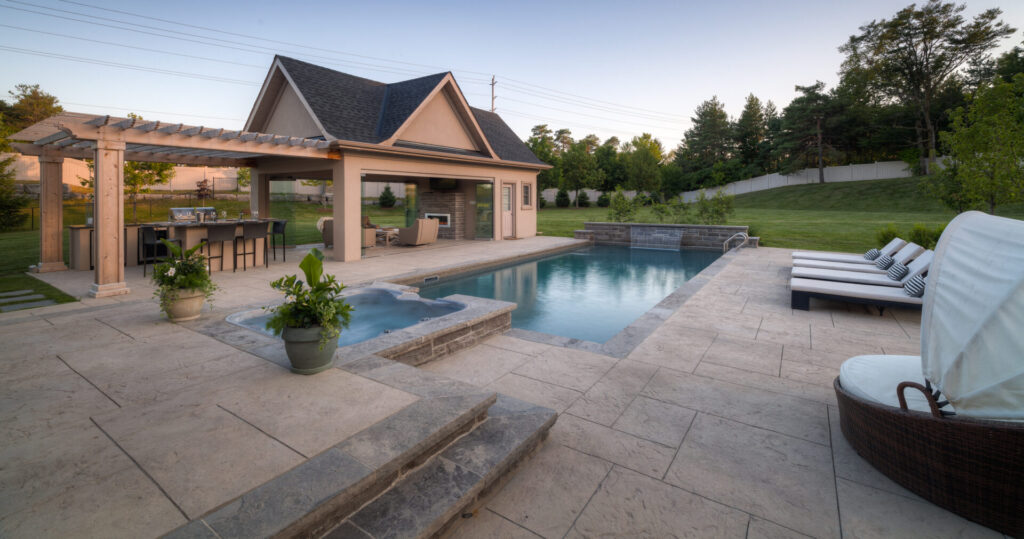 Luxurious backyard with a swimming pool, hot tub, pergola, outdoor kitchen, lounge area, and neatly trimmed grass surrounded by a tall privacy fence at dusk.