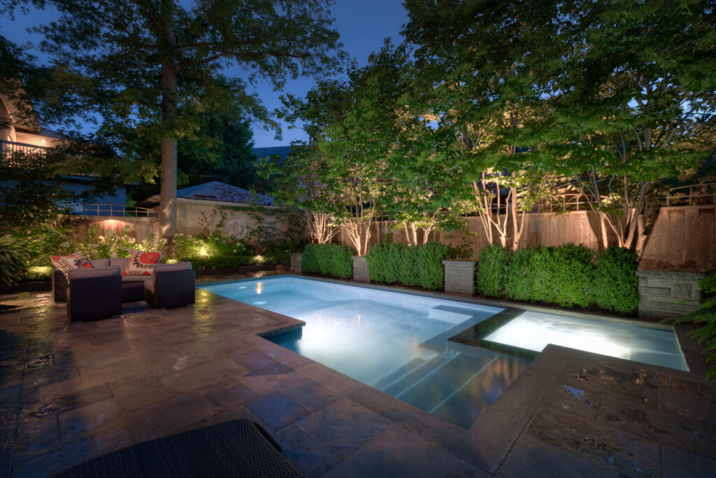 An evening shot of a serene backyard with a lit rectangular pool, stone patio, outdoor furniture, trees, bushes, and a wooden fence for privacy.