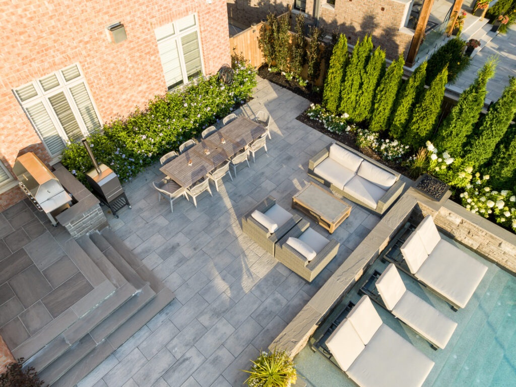 An aerial view of a neatly organized outdoor patio with furniture, a dining area, a grill, poolside loungers, and lush greenery by a brick building.