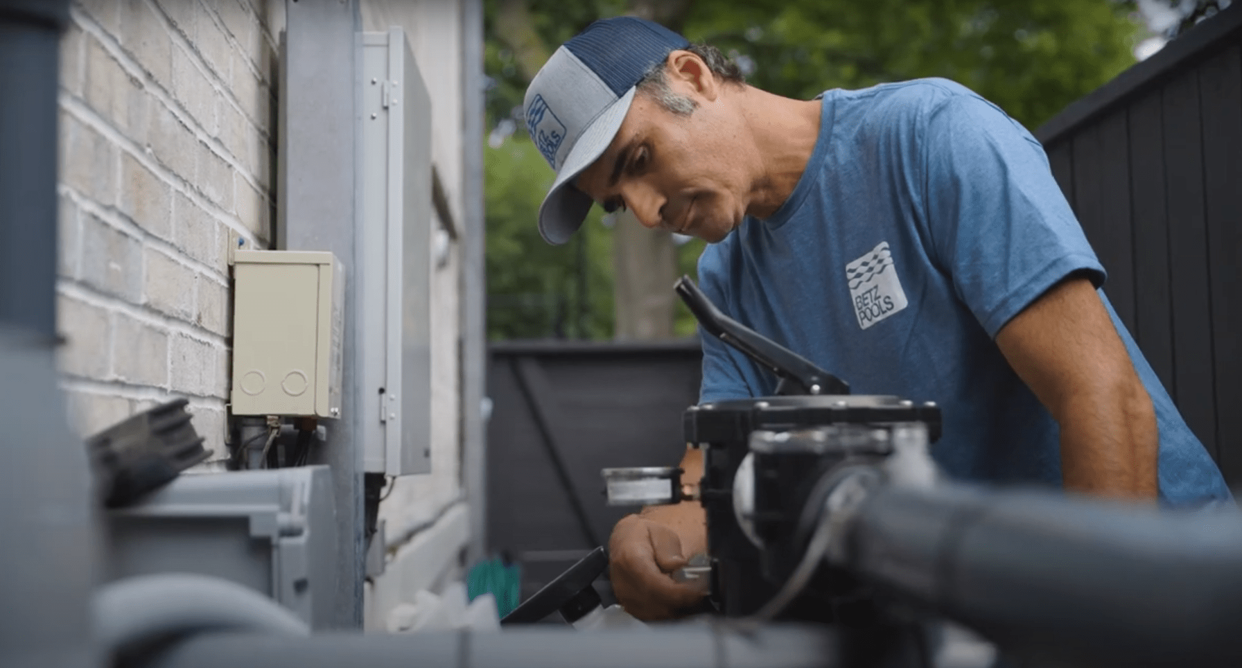 A person wearing a baseball cap and a blue t-shirt is focusing on repair work, possibly on HVAC equipment, outside against a brick wall.