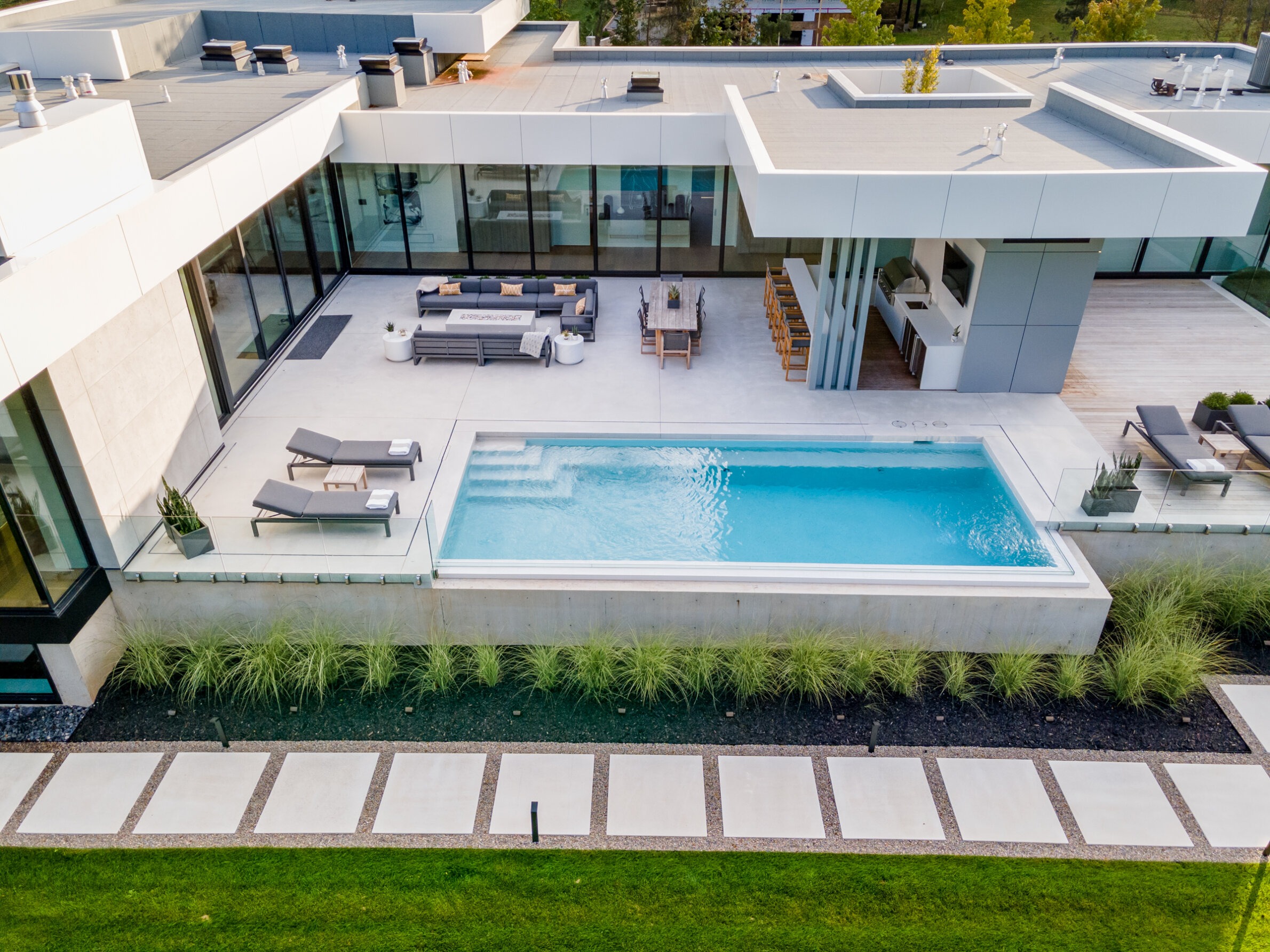 A modern house with flat roofs, expansive windows, an outdoor pool, patio furniture, and carefully landscaped garden with a path of stepping stones.