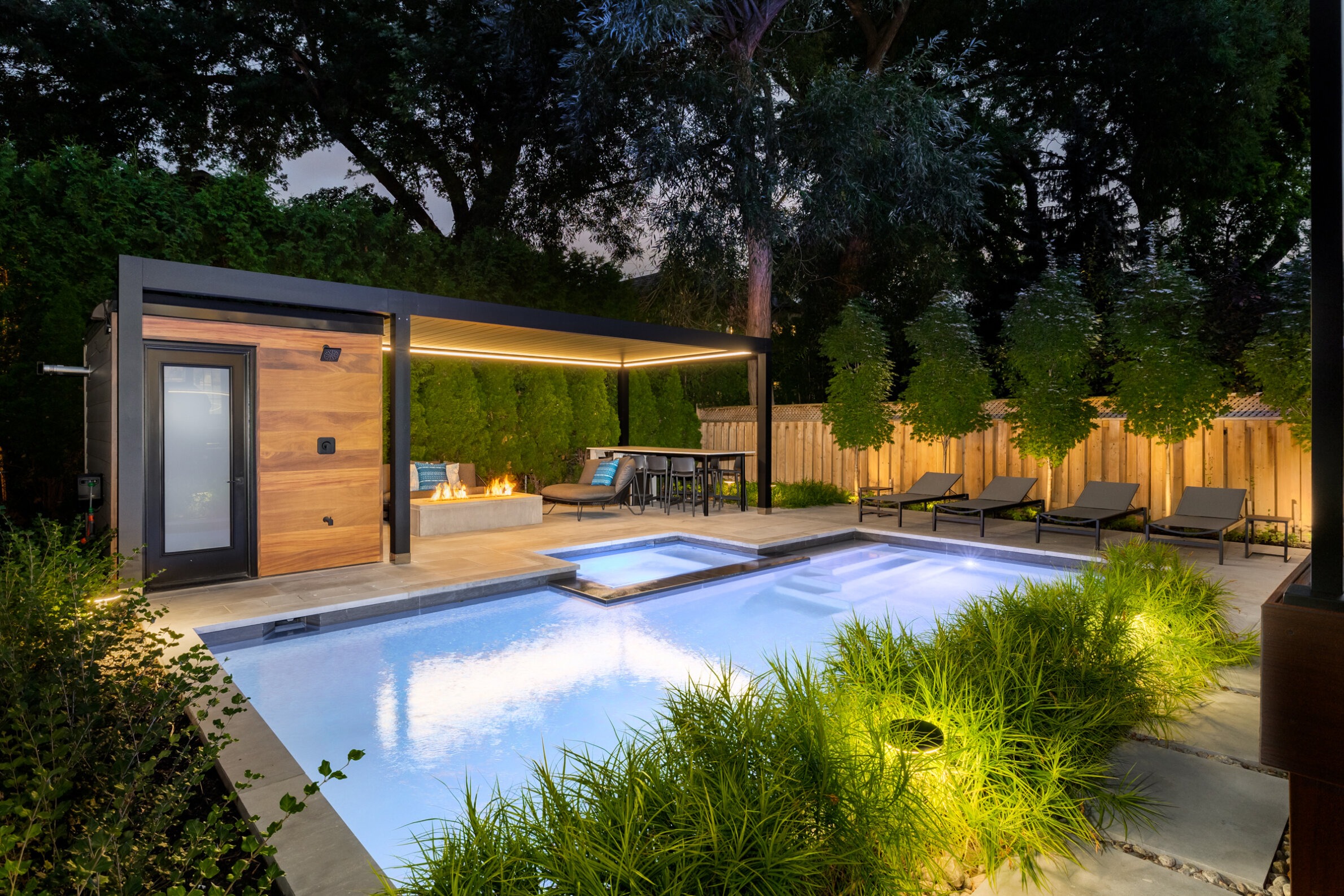 Modern backyard at dusk featuring a swimming pool, hot tub, outdoor seating, fire pit, chaise lounges, lighting, and lush greenery.