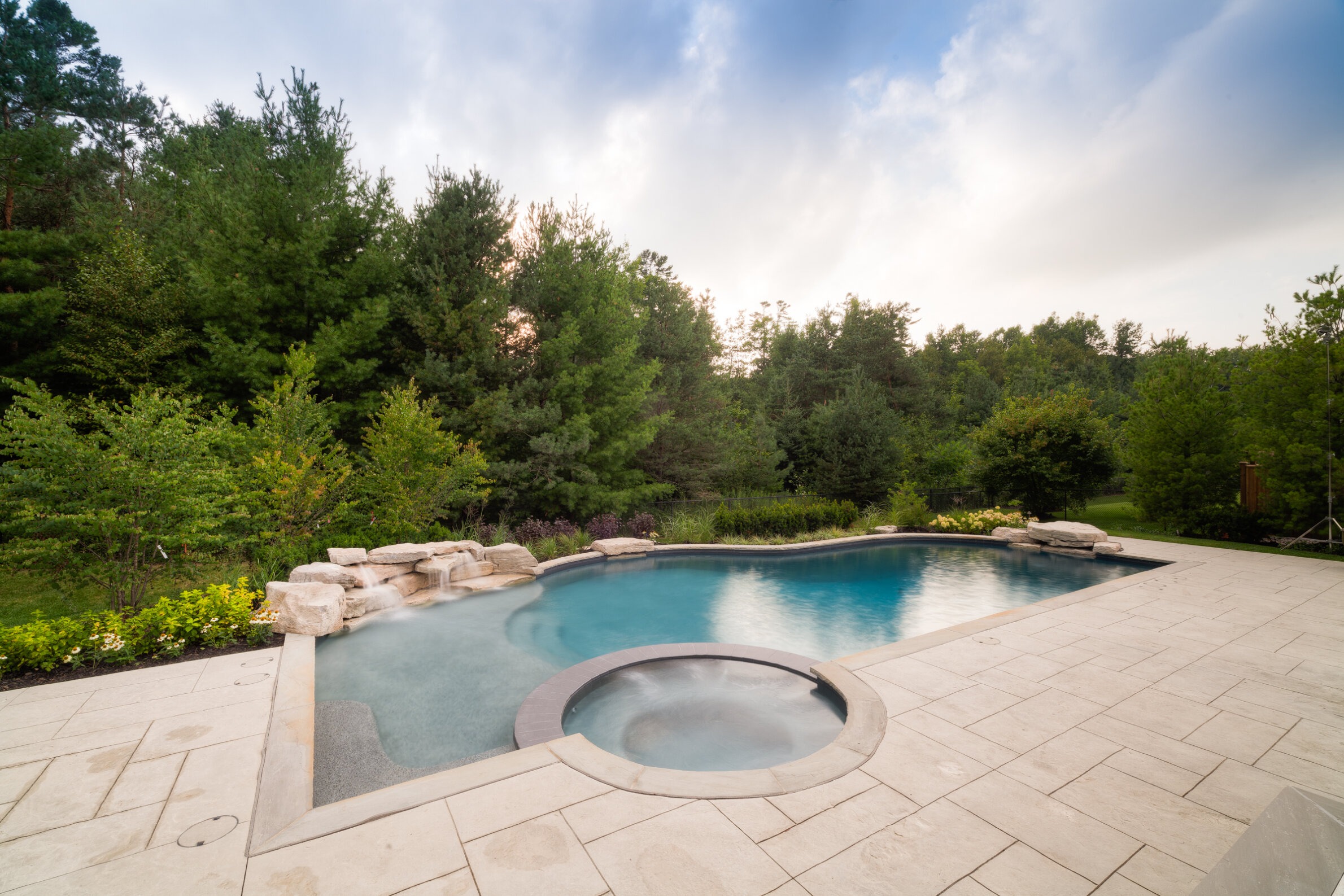 The image shows a backyard with a curvy swimming pool and a hot tub, surrounded by a stone patio and lush green trees under a cloudy sky.