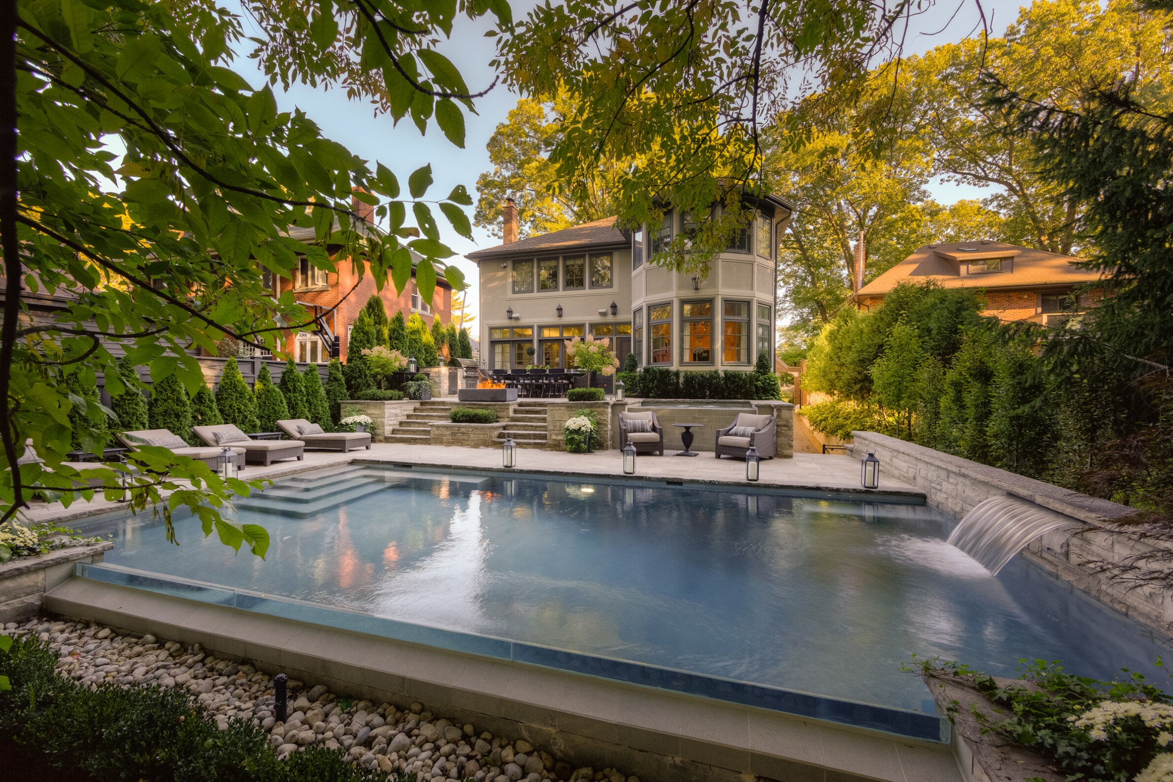 An elegant backyard with a large swimming pool, surrounded by manicured greenery, comfortable outdoor furniture, and a two-story house with large windows.