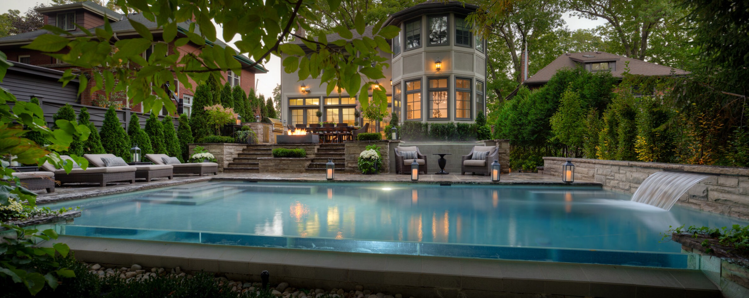 Luxurious backyard at dusk with an illuminated pool, lounge chairs, lush greenery, a cozy fire pit area, and an elegant home in the background.