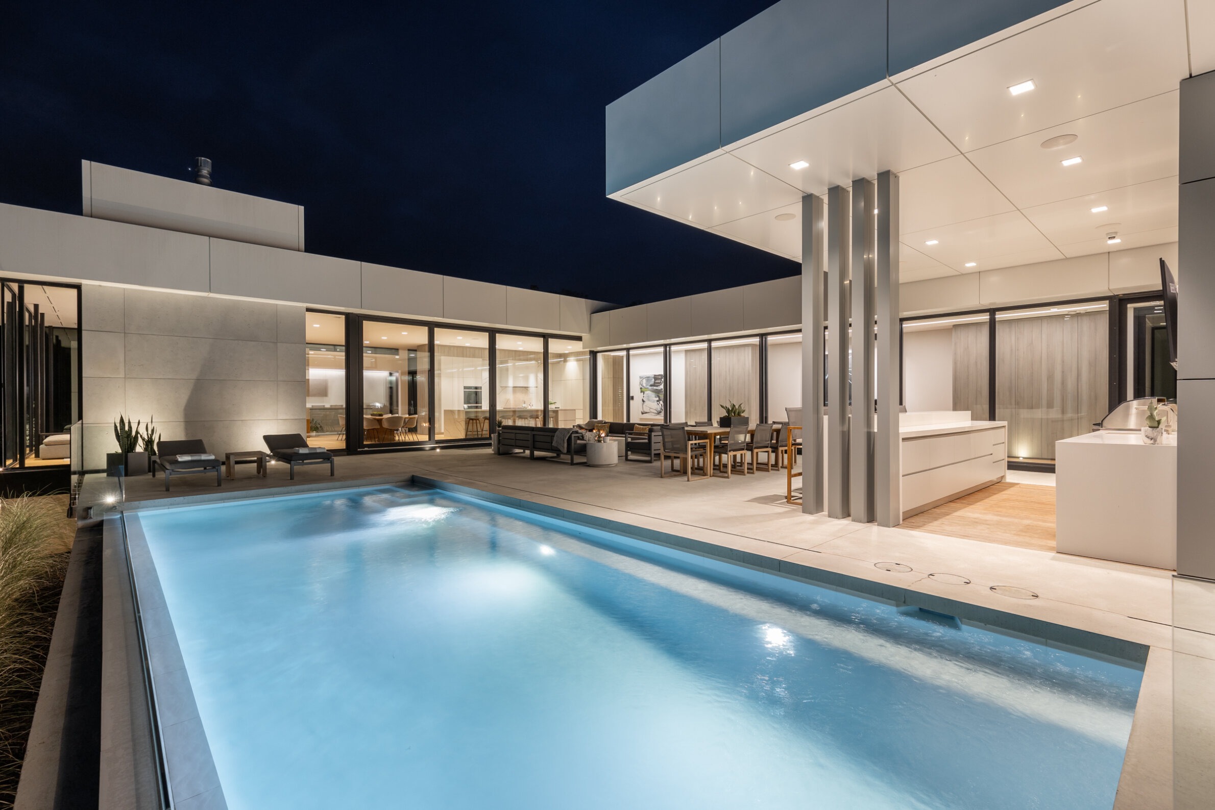 A modern house with an outdoor pool lit at night, sleek architecture, large windows, and a comfortable patio area with furniture and lighting.