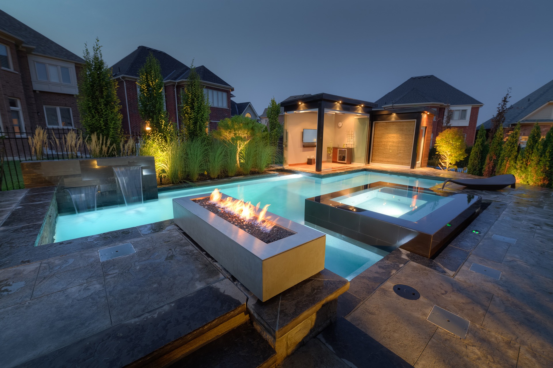 Luxurious backyard at dusk featuring a swimming pool with waterfall, a hot tub, a lit fire feature, landscaped garden, and a modern pool house.