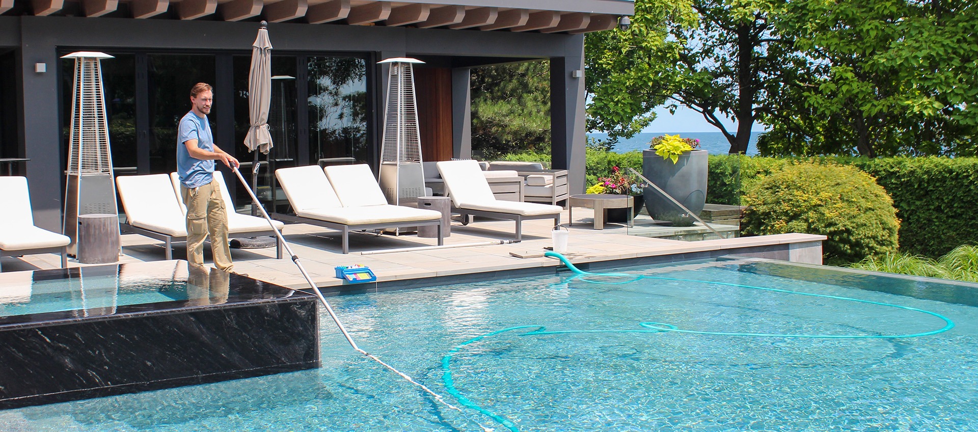 A person is cleaning a luxurious infinity pool with a skimmer net, surrounded by sun loungers, in a scenic outdoor setting with lush greenery.