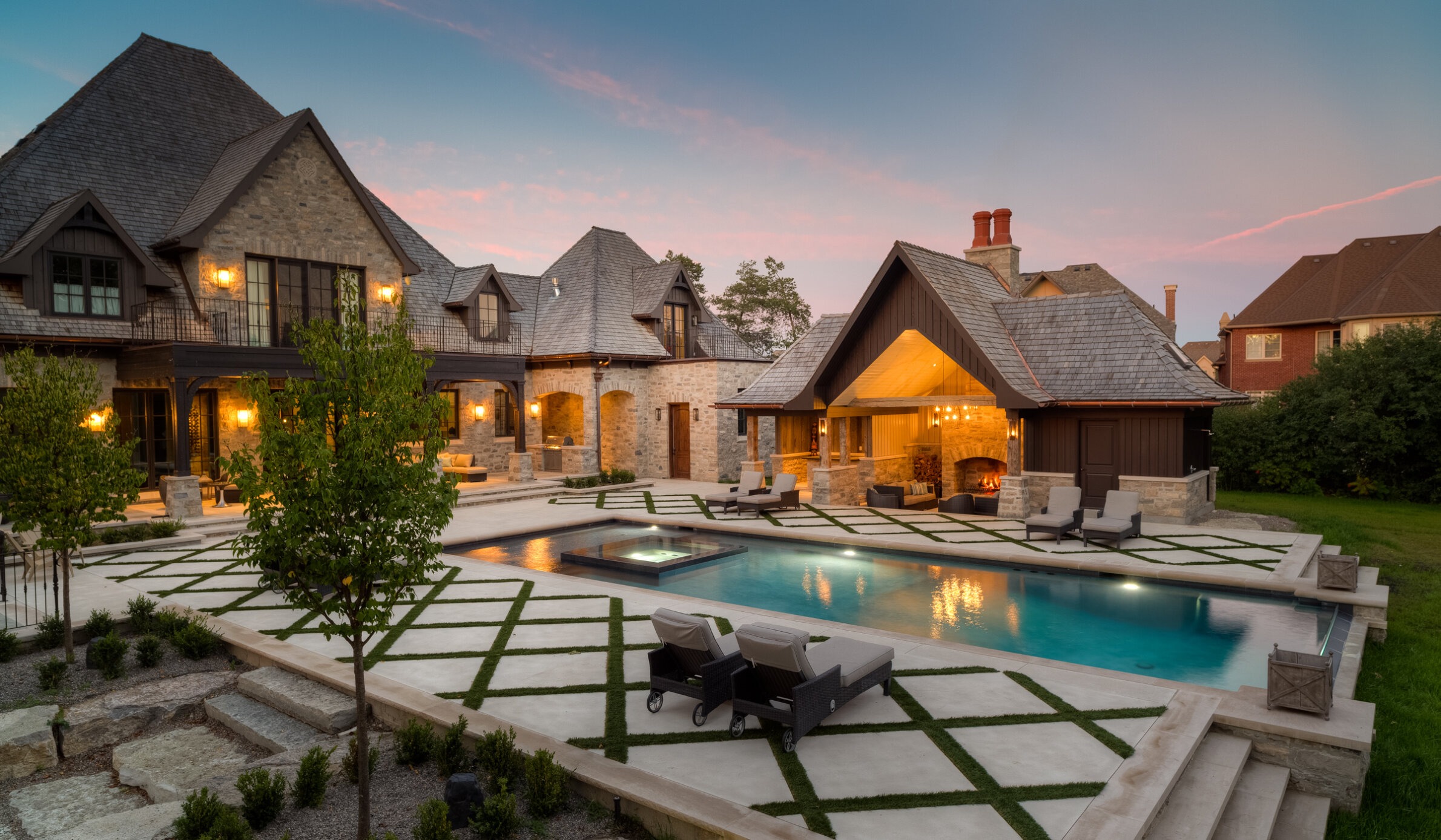 Luxurious backyard with a pool, lounging chairs, lush landscaping, and a stone house with lit windows at dusk under a soft pink sky.