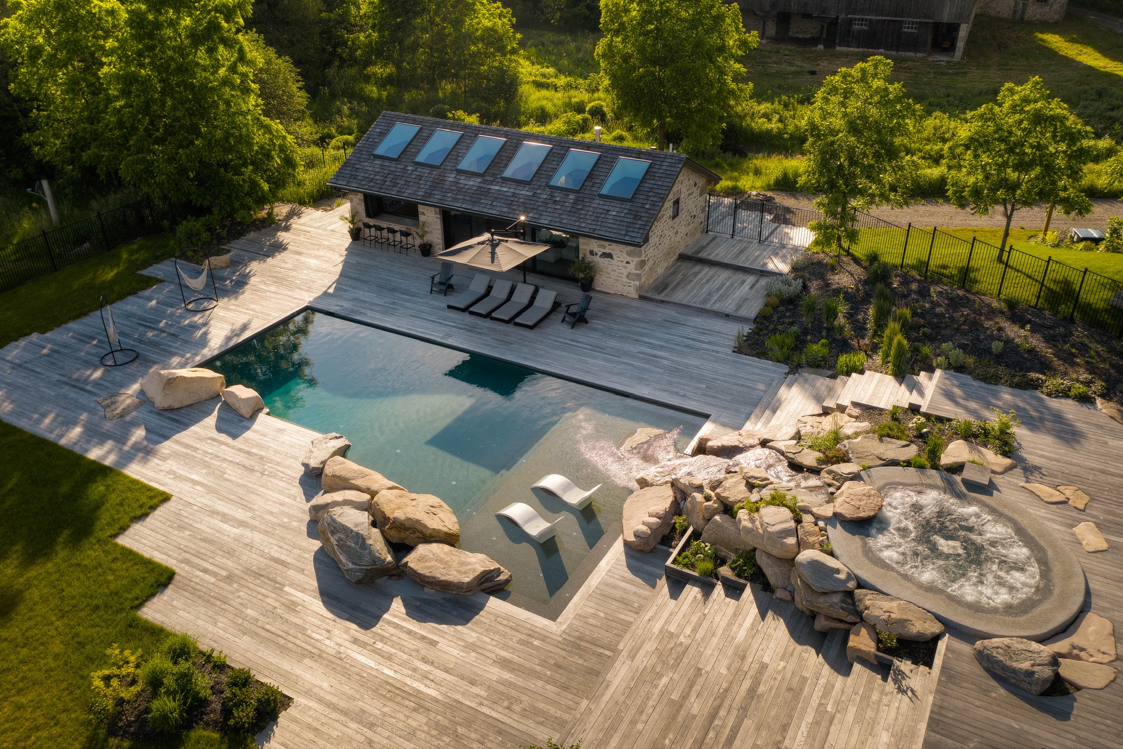 A modern backyard with a rectangular pool, hot tub, lounging chairs, wooden deck, and a stone house surrounded by lush greenery and fencing.