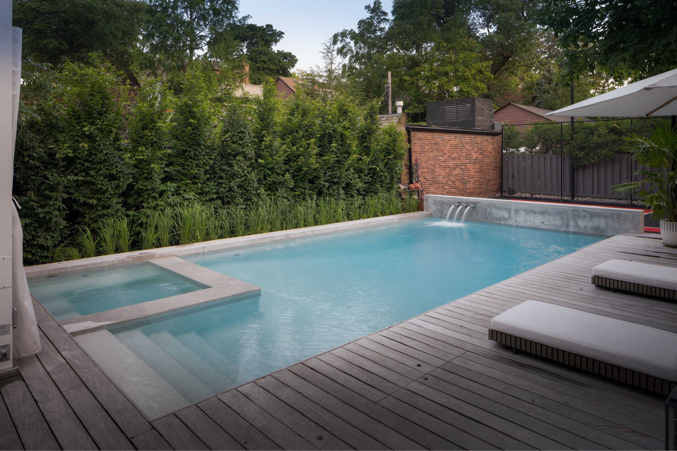 A serene backyard setting with a large swimming pool, surrounded by wooden decking and lush greenery, featuring a hot tub and recliners.