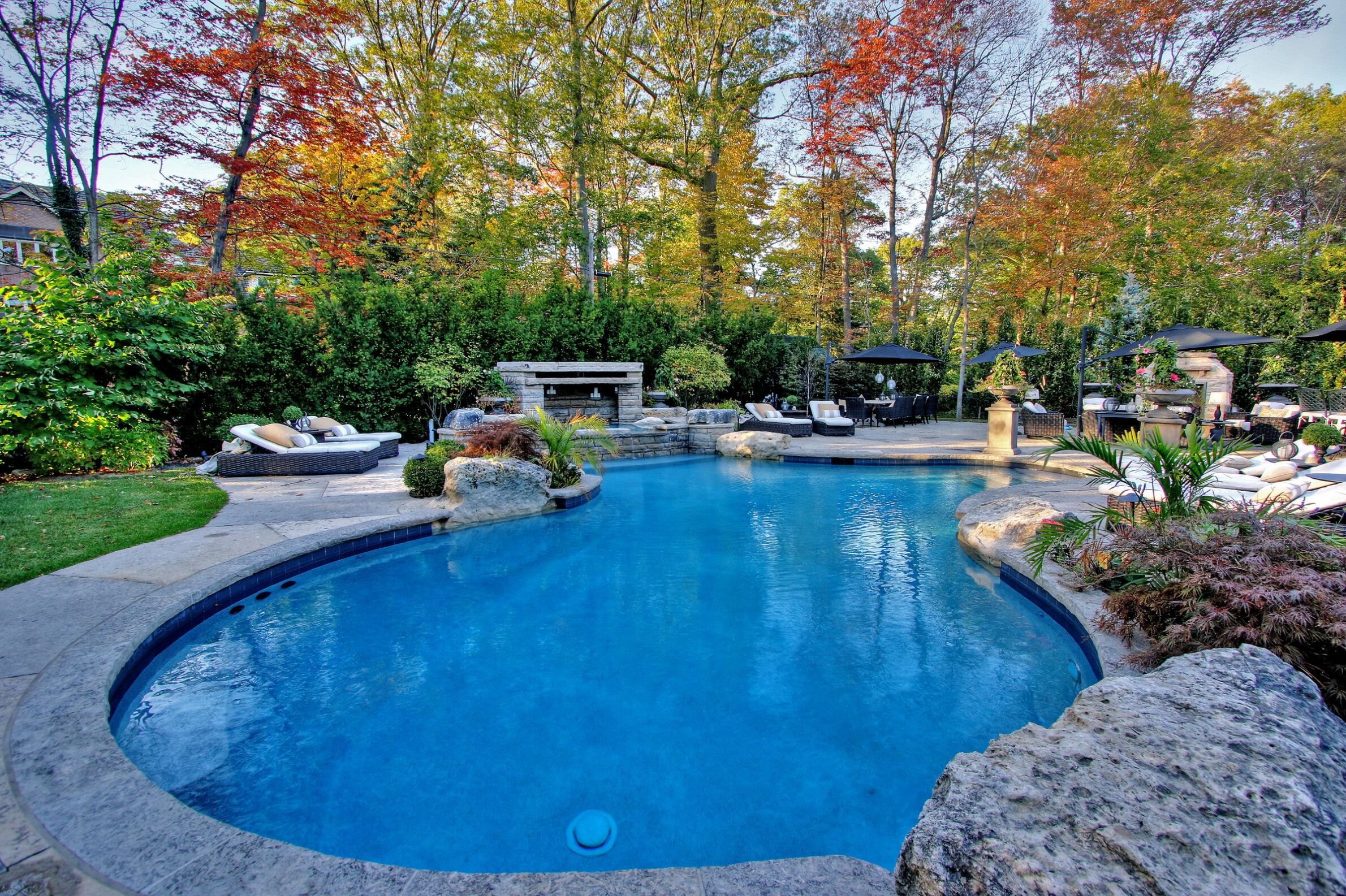 An inviting backyard with a curvy swimming pool, surrounded by autumn-colored trees, patio furniture, outdoor fireplace, and lush greenery under clear skies.