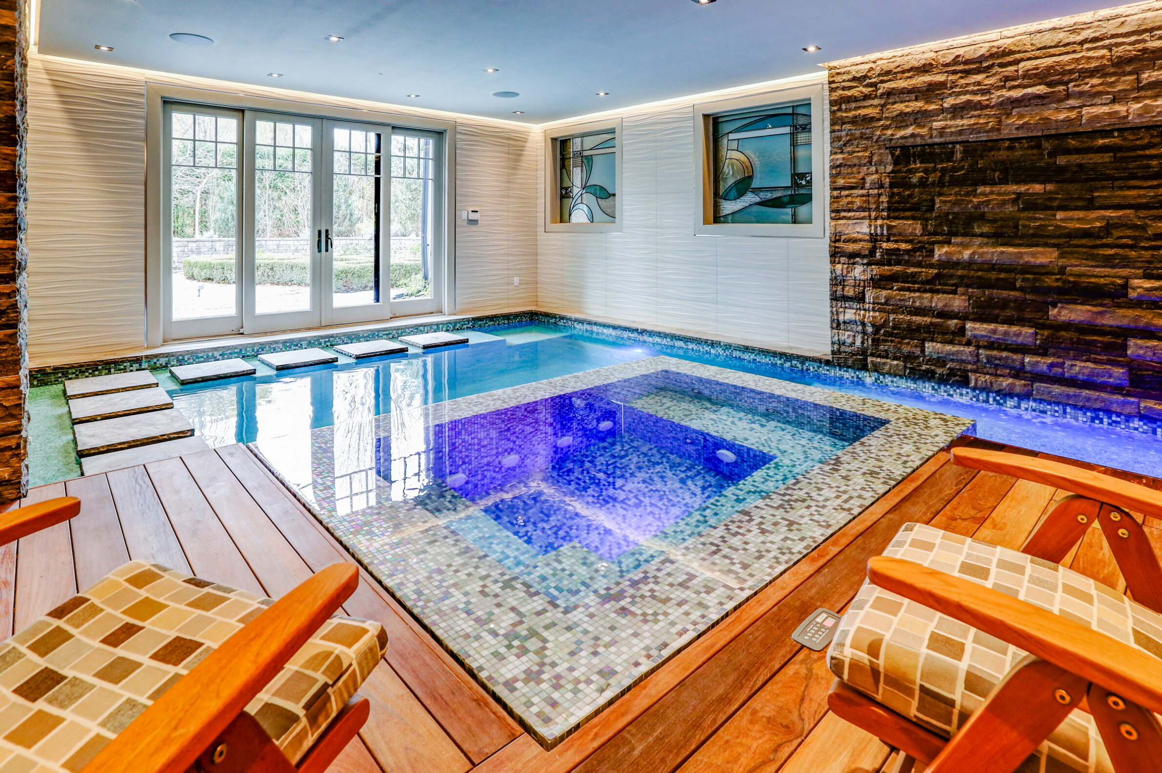 An indoor swimming pool with mosaic tiles, wooden decking, and loungers. Natural light streams in; a stone feature wall and art add elegance.