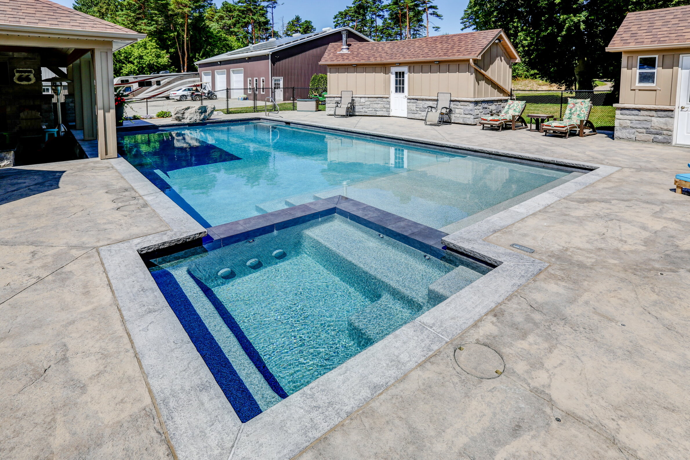 This image features a clear blue rectangular swimming pool with an integrated hot tub, surrounded by a concrete deck and buildings, on a sunny day.