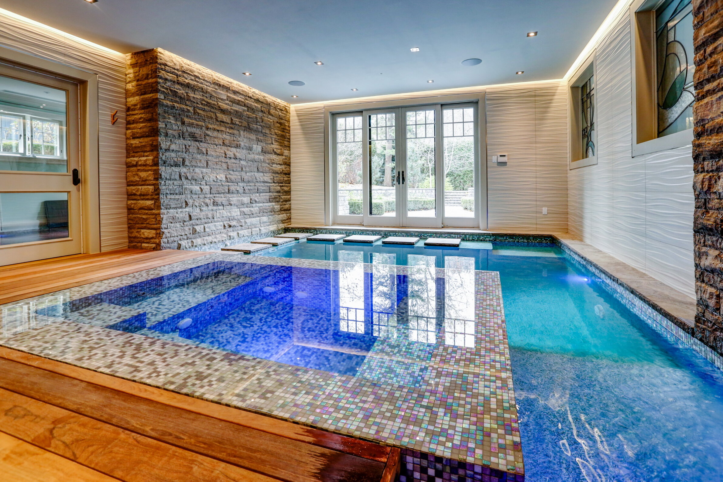 Indoor swimming pool with blue mosaic tiles, wooden deck, natural light through large windows, and a stone accent wall on one side.