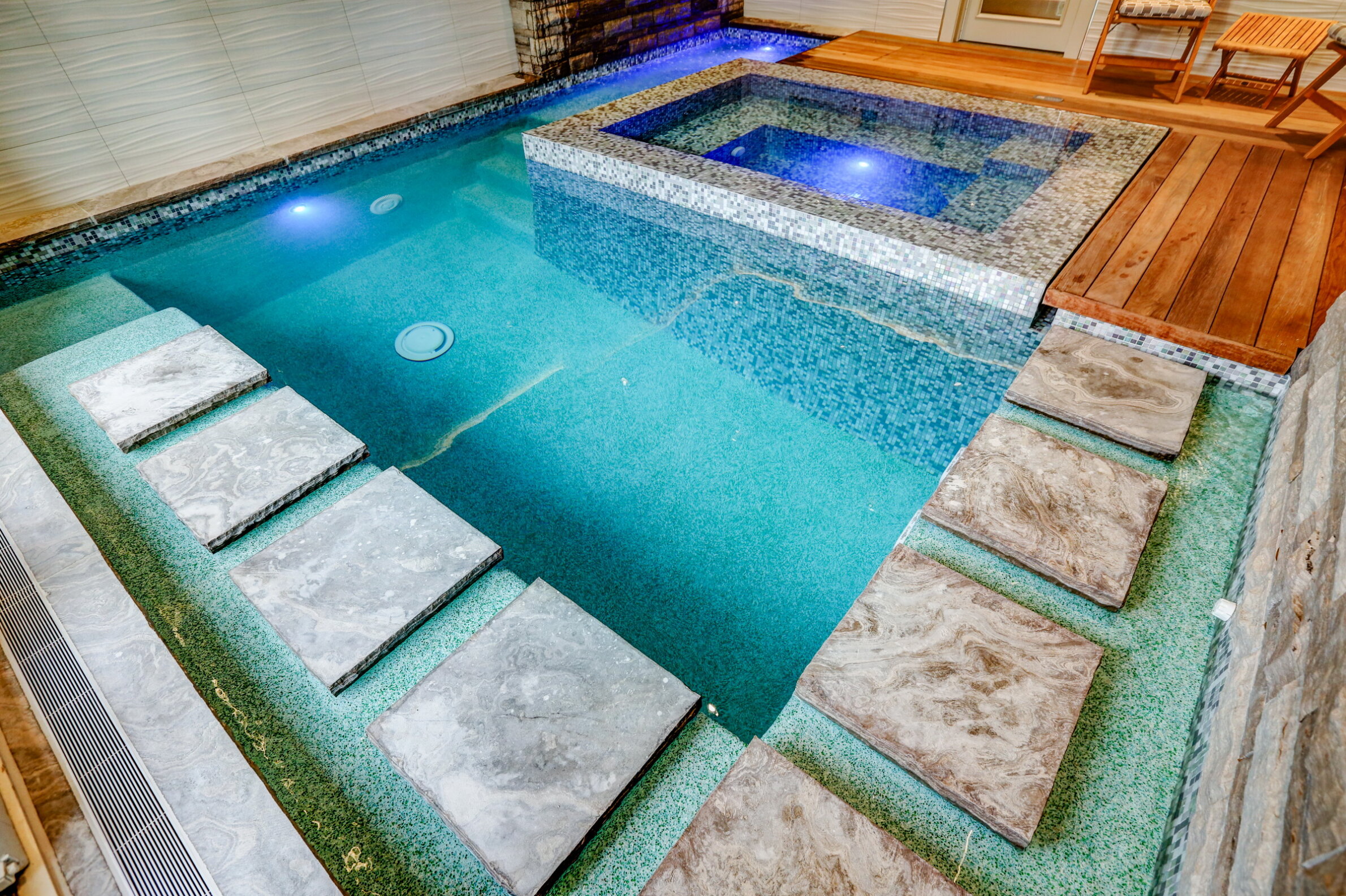 Indoor pool with a sleek design featuring stone steps, a blue mosaic tile spa section, ambient lighting, wooden deck, and a stone wall.