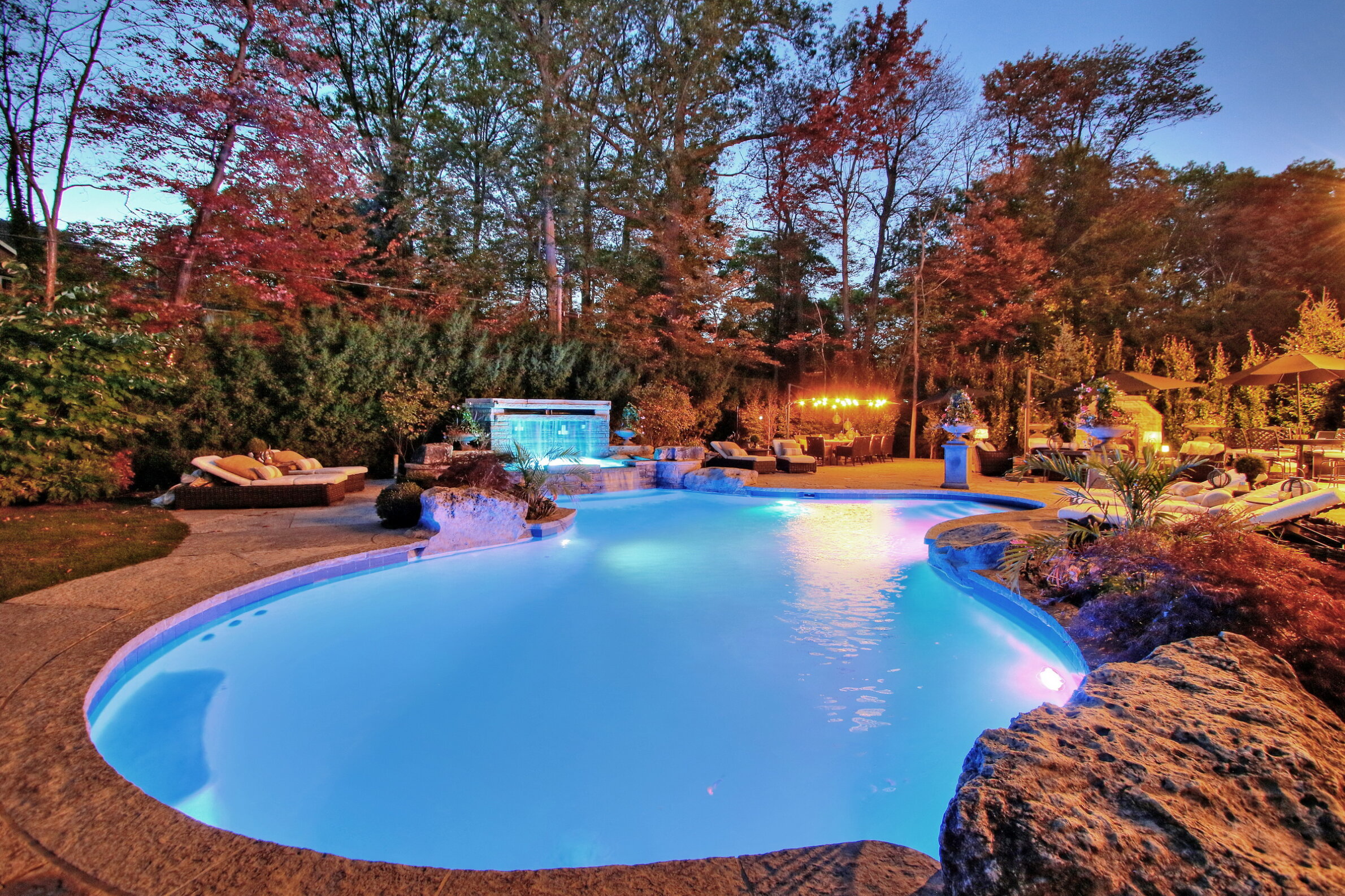 An outdoor swimming pool with blue lighting at dusk, surrounded by autumn trees, cozy lounge areas, and a water feature in a backyard setting.